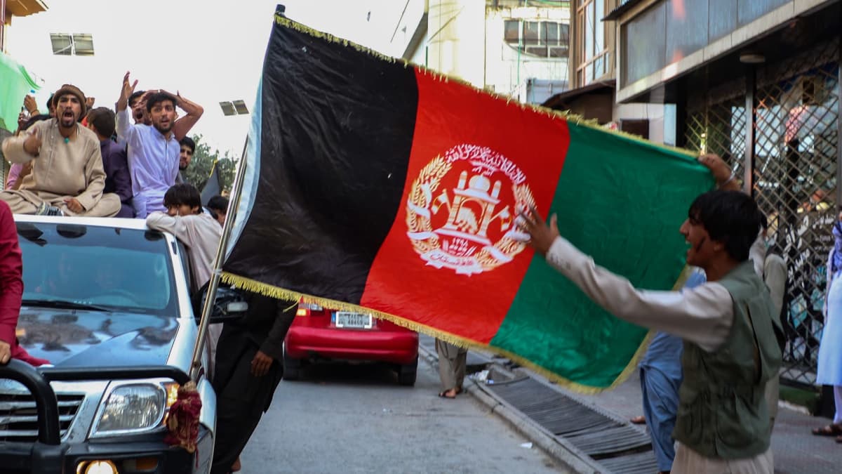 Mies pitelee Afganistanin lippua. Taustalla auto täynnä miehiä.