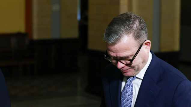 A man in a blue suit in a dimly lit hall with his head hanging down.