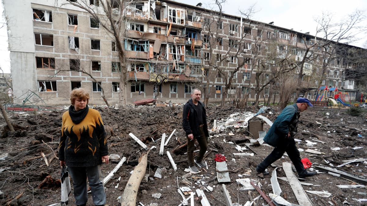 Kolme henkilöä kävelee pommituksissa tuhoutuneen kerrostalon pihalla.