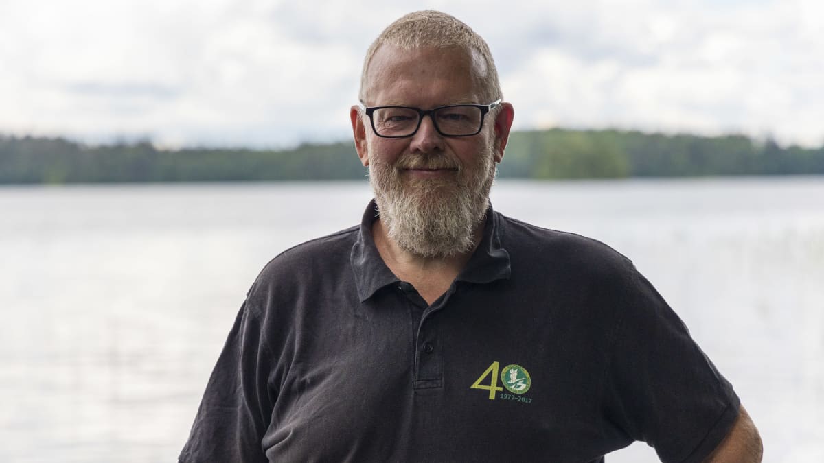 Suomen Luonnonvalokuvaajien puheenjohtaja Kari Wallgren seisoo järven rannalla katsoo kameraan. 