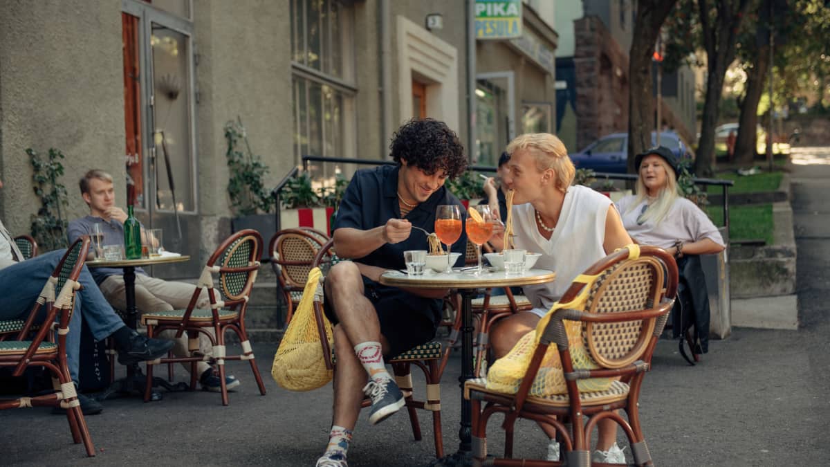Två unga män sitter och äter spaghetti på en uteservering i stadsmiljö.