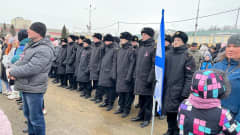 Joukko sotilaita seisoo rivissä aukiolla väkijoukon keskellä. Yhdellä sotilaalla on valko-sininen lippu kädessään.