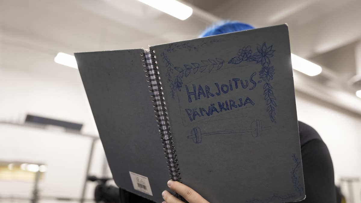 Anonyymi henkilö täyttää harjoituspäiväkirjaa. Päiväkirjan kannessa lukee harjoituspäiväkirja ja siihen on piirretty painnonnosto tanko painoineen.