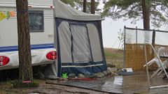 Husvagn med en altan ute gjord av trädäck. Plaststolar, tält som förstorar husvagnen och bord. Regnigt. Man ser strand och hav i bakgrunden. Silversand camping i Hangö.