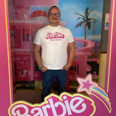 Vaalea mies valkoisessa Barbie-paidassa seisomassa pahvisessa Barbie-elokuvan kuvakehysrekviisitassa.