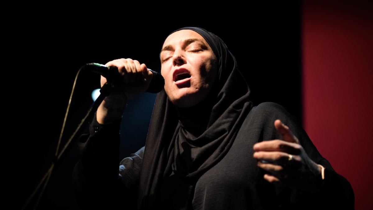 Sinead O’Connor laulaa ja pitää mikistä kiini. Hänellä on musta huntu yllään.