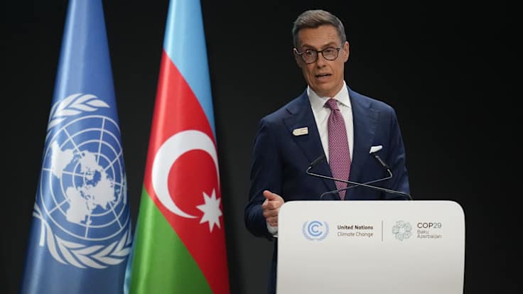 Man in a dark suit, white shirt and red tie, speaking at a podium with the flags of the UN and Azerbaijan in the background.