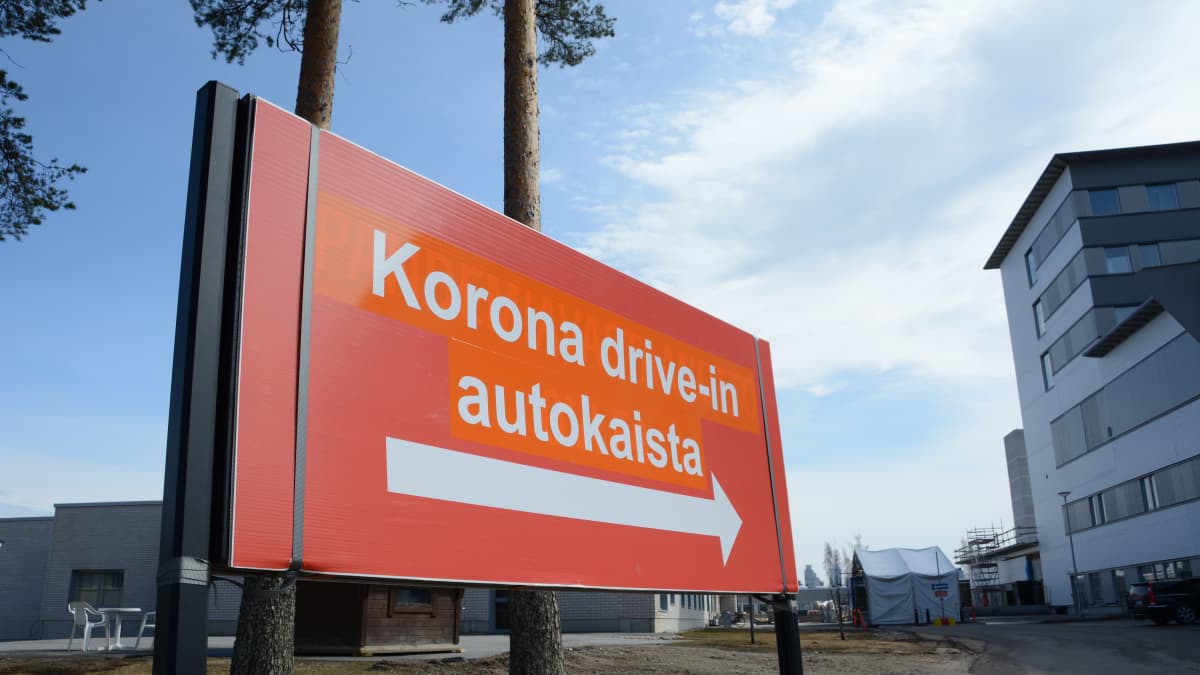Iso oranssi kyltti Koronatestauksesta Kajaanin keskussairaalan pihassa. Taka-alalla näkyy teltta, jossa drive-in -testaukset suoritetaan.
