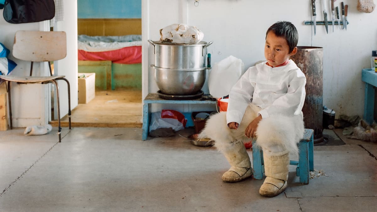 Jääkarhuhousuihin pukeutunut nuori lapsi istuu sinisellä tuolilla huoneessa. Takana näkyy keittovälineitä ja vaatteita.