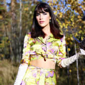 Romy Räihälä seisoo metsässä vihreässä, kaksiosaisessa asussa, jossa on pinkkejä kukkia. Hänellä on 60-luvun tyylinen meikki. 