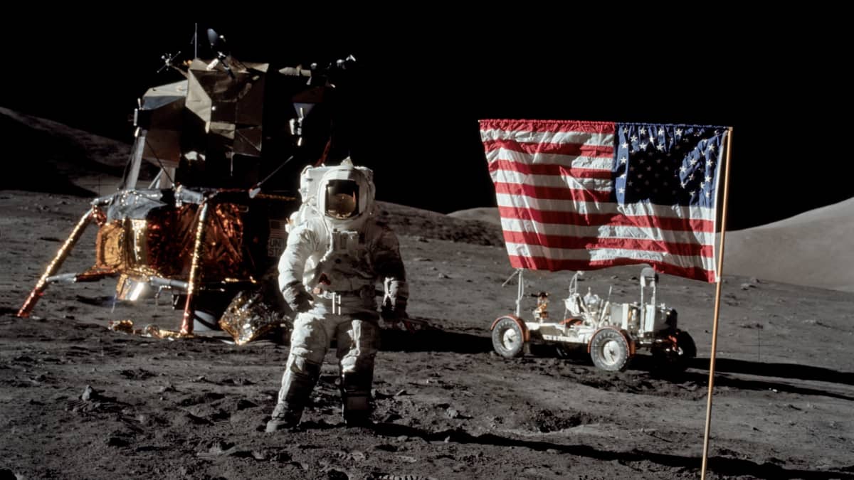 Astronautti seisoo Kuussa Yhdysvaltain lipun vieressä.