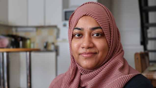 En småleende kvinna med rosa sjal runt huvud och hals fotograferad i ett kök.