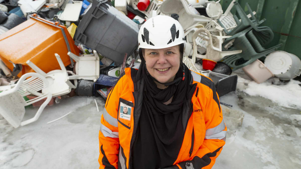Mari Juntunen seisoo muovitavaraläjän edustalla, katsoo ja hymyilee ylös kameraan suojakypärä päässään ja oranssi turvatakki päällään.
