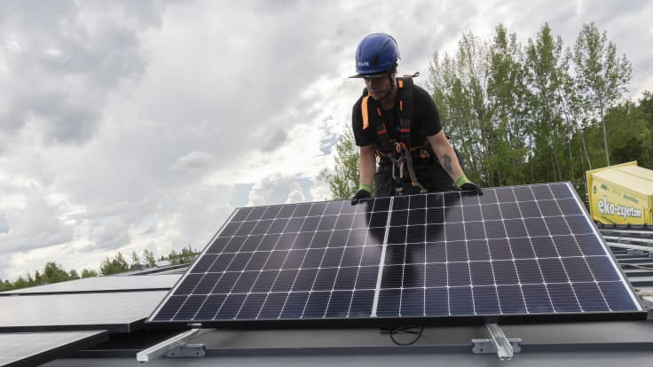 Aurinkopaaneelien asentaja Juha Matilainen asettelee suurta aurinkopaneelia kiskoille katolla.