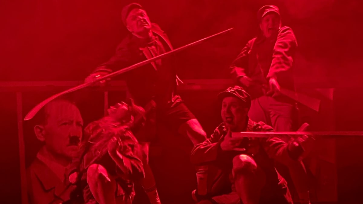 Punaisen valon sävyttämä näyttämökuva. Näyttämöllä hidastetuissa asennoissa neljä näyttelijää sotilaspuvuissa epämääräisenaä rykelmänä, käsissään pyssyä muistuttavat jääkiekkomailat. Taustalla Hitlerin pahvikuva.