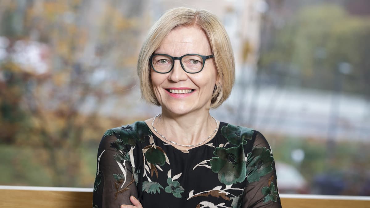 kasvatustieteen professori Anna Rönkä istuu ikkunan edessä kädet puuskassa ja hymyilee kameralle.