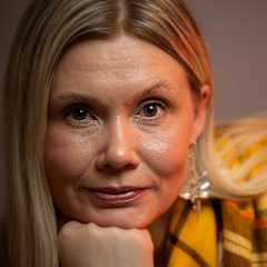 Kuvassa on henkilö, jolla on pitkät vaaleat hiukset. Hänellä on yllään keltainen huivi. Tausta on yksivärinen ja hillitty, ja valaistus on lämmin. 