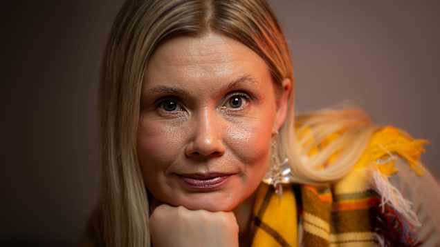 Kuvassa on henkilö, jolla on pitkät vaaleat hiukset. Hänellä on yllään keltainen huivi. Tausta on yksivärinen ja hillitty, ja valaistus on lämmin. 