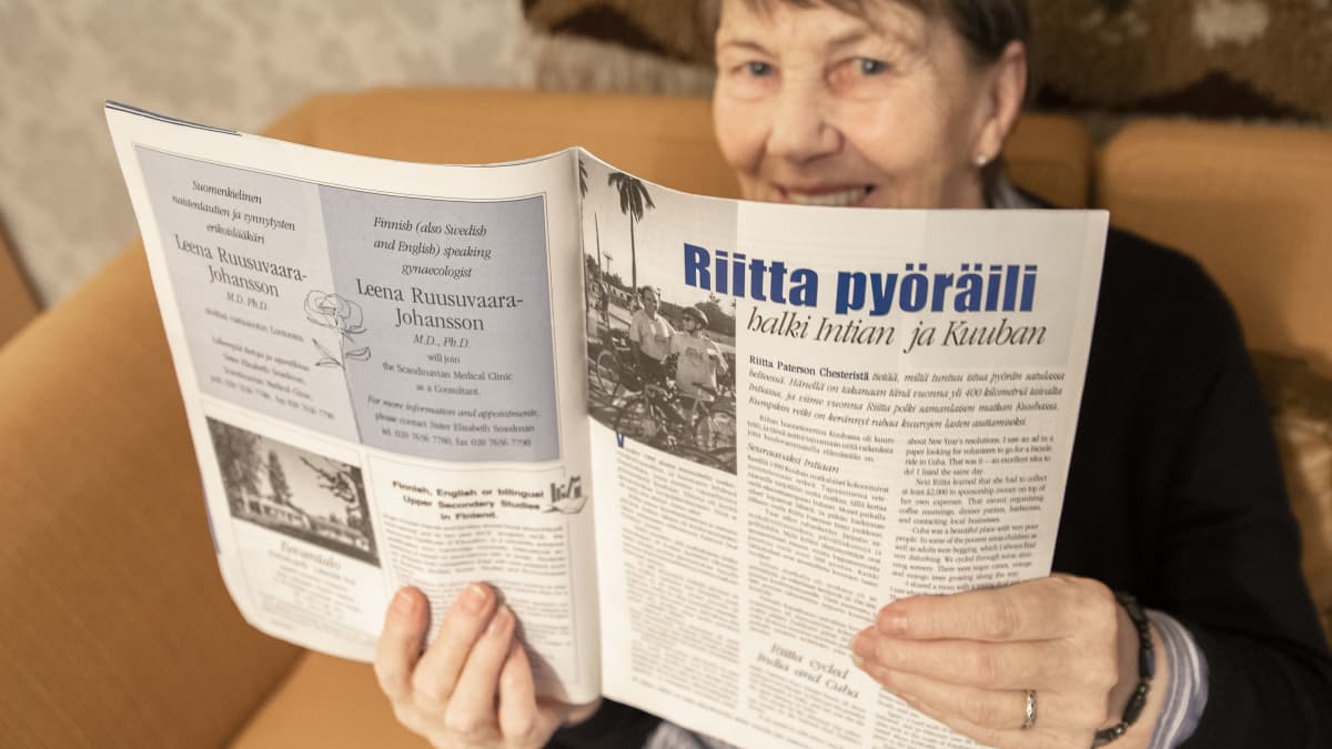 80 vuotta täyttävä Riitta Paterson istuu sohvalla ja näyttää lehteä, jossa kerrotaan hänen tekemistään pyöräilymatkoista ulkomailla