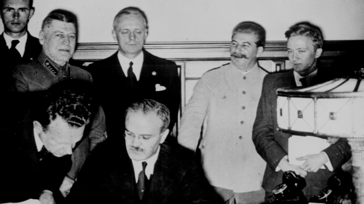 Molotov istuu pöydän ääressä allekirjoittamassa. Hänen takanaan seisoo Ribbentrop, vakavana. Stalin hymyilee vieressä univormussaan.