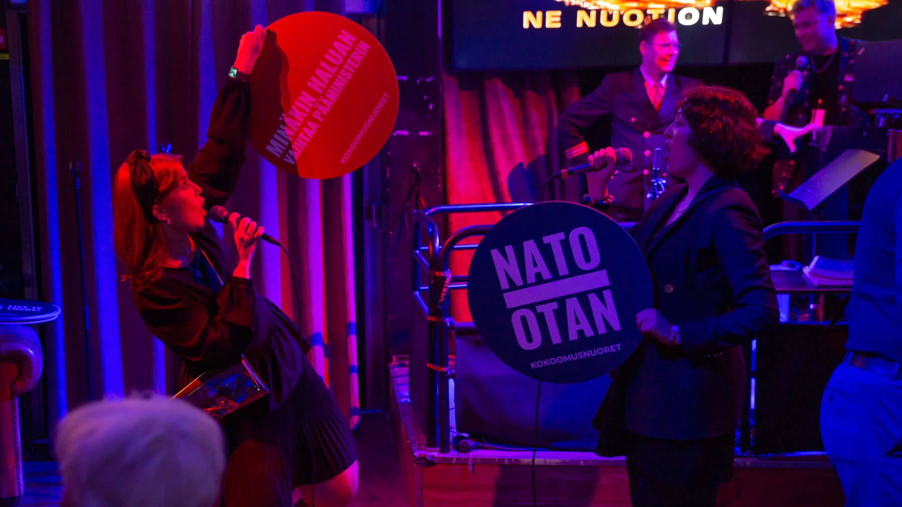 Kansanedustajaehdokas Teresa Rautapää Lahdesta ja Päivi Lindqvist Hämeenlinnasta laulavat Kokoomuksen risteilyllä karaokessa. Molemmilla on käsissään kokoomusnuorten iskulausekylttejä joissa lukee: "Minäkin haluan vaihtaa pääminiserin" ja "NATO - OTAN".