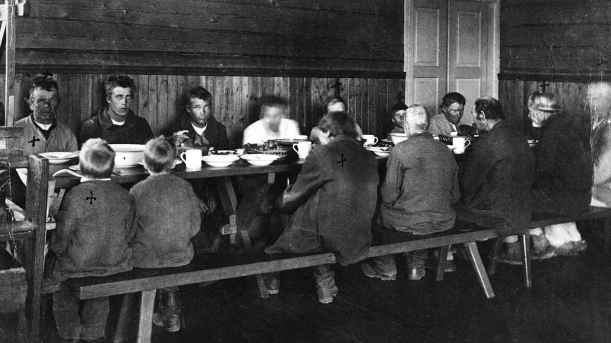 Köyhiä ihmisiä ruokapöydän ääressä mustavalkoisessa kuvassa. 