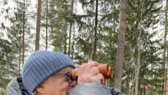 Mustaan toppatakkiin ja siniseen pipoon pukeutunut keski-ikäinen mies katsoo kiikarilla metsään.