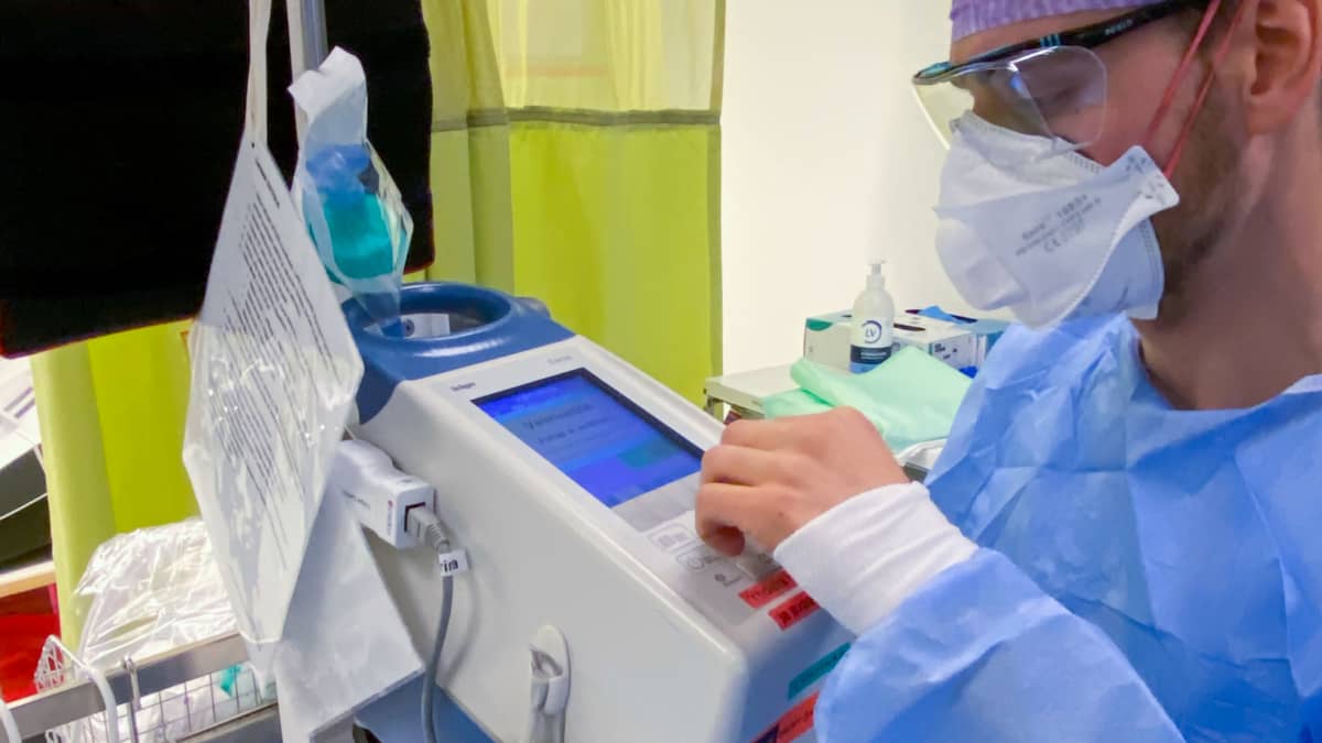 Anestesialääkäri sinisessä suoja-asussa tarkkailemassa hengityskonetta sairaalan teho-osastolla.