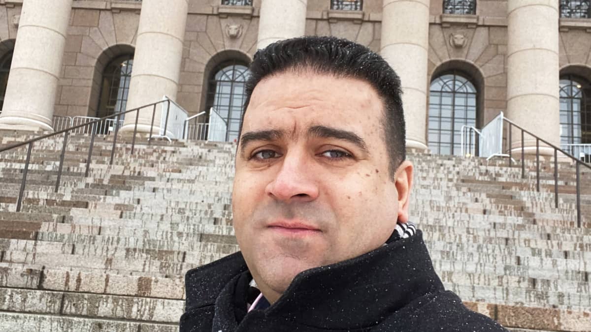 Selfie, jossa tummahiuksinen mies seisoo eduskuntatalon portailla ja taustalla näkyy eduskuntatalon julkisivu.