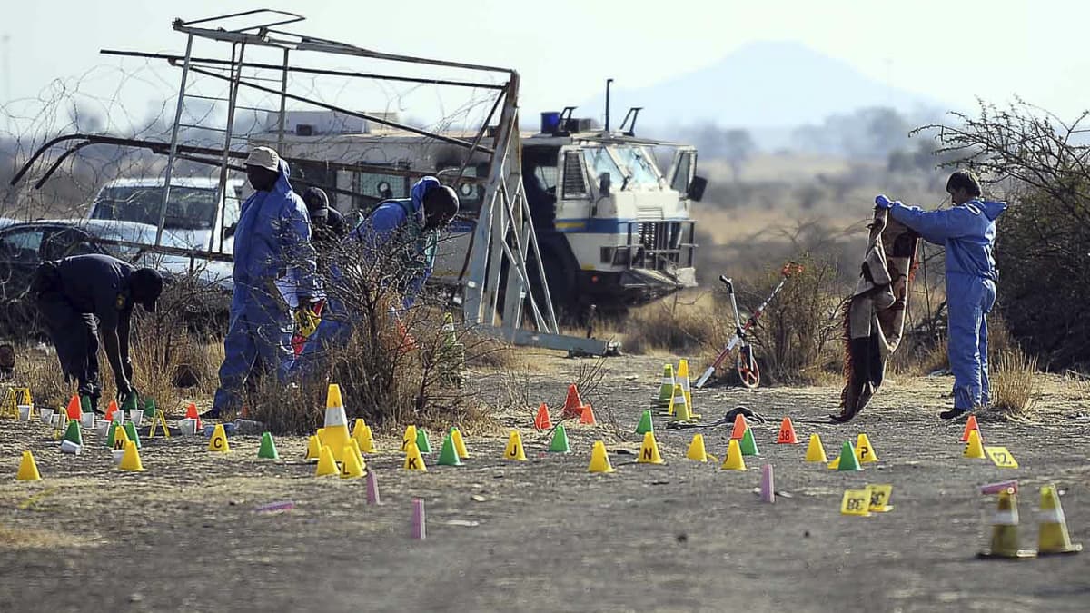 Några personer i blå overaller utför undersökningar efter massakern i Marikana. Gula, gröna och orange markeringar på marken. Några fordon i bakgrunden. 