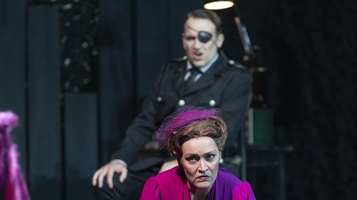 Fricka (Niina Keitel) on polvillaan näyttämöllä, hänen takanaan näkyy silmälapulla varustettu Wotan (Tommi Hakala).
