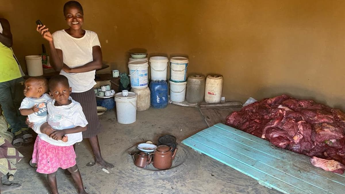 Afrikkalainen äiti ja kaksi lasta kodissaan, jossa pöydällä norsun lihaa.