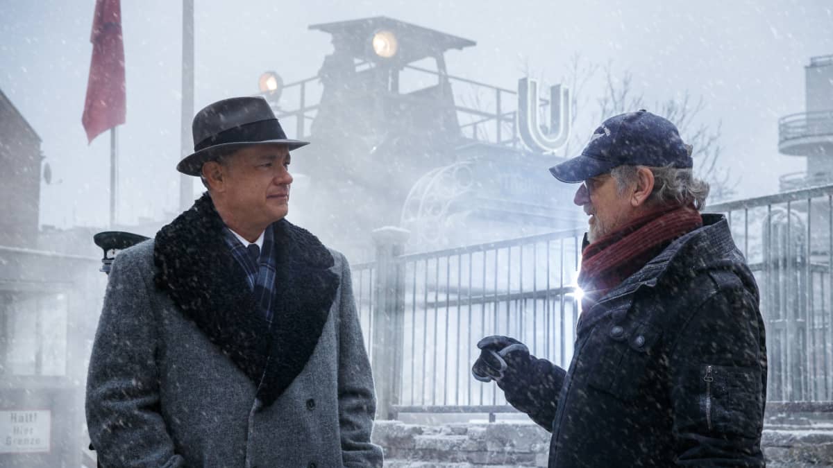 Villakangastakkiin ja fedoraan pukeutunut mies keskustelee lippalakkipäisen, harmaakiuksisen miehen kanssa lumisateessa.