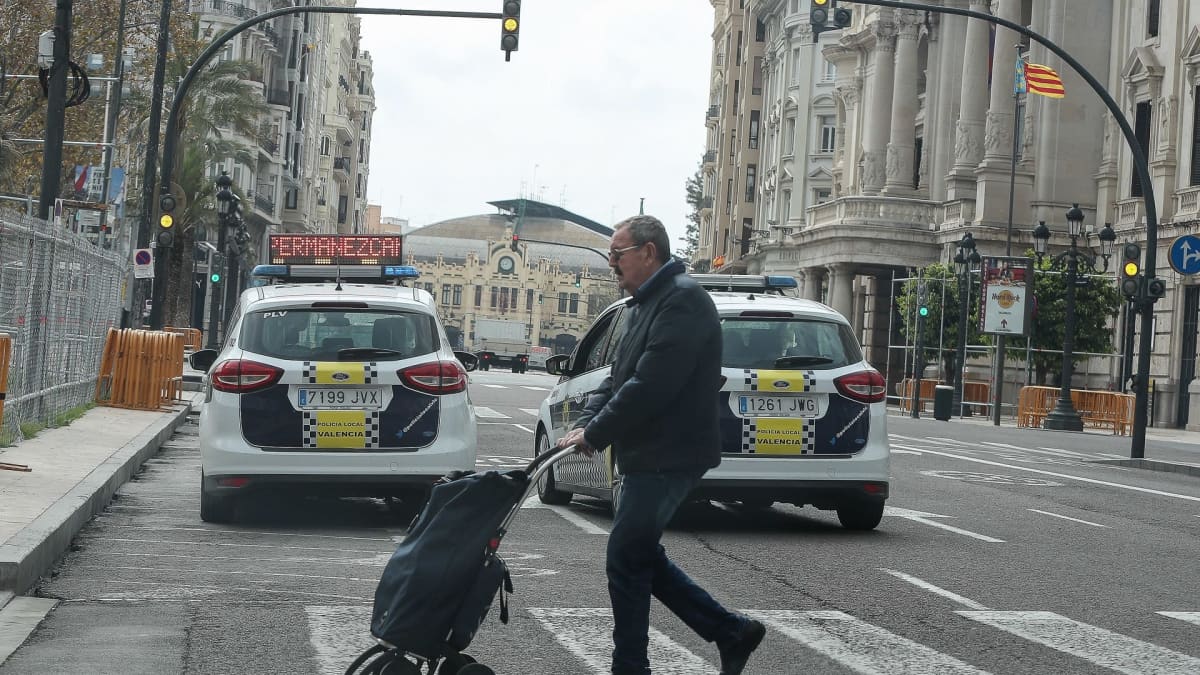 Mies kulkee suojatiellä vedettävän ostoslaukun kanssa. Muita ihmisiä ei näy, vain poliisiautoja.
