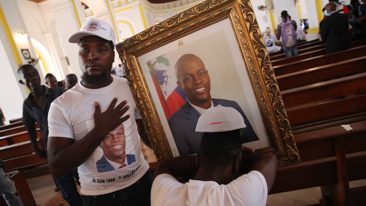 Mies pitelee kirkossa presidentti Jovenel Moisea esittävää taulua. Presidentin kuva on myös hänen paidassaan. Toinen mies nojaa päätään käsiinsä.