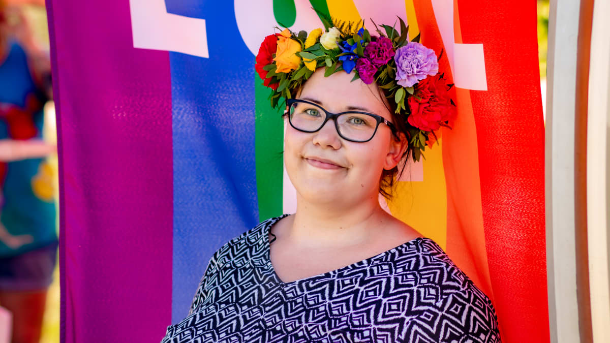 Silmälasipäinen nuori nainen kukkaseppele päässään hymyilemässä kameralle. Taustalla Pride-lippu.