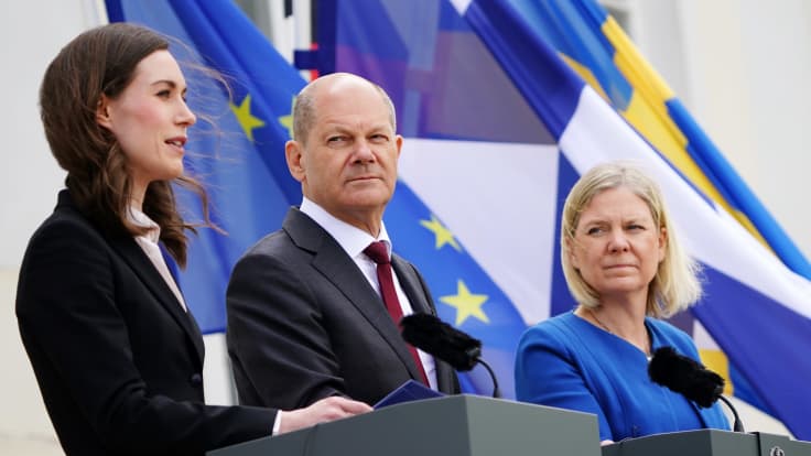 Sanna Marin, Olaf Scholz och magdalena Andersson håller presskonferens bakom var sitt podie i blåsiga förhållanden. I bakgrunden ländernas flaggor.