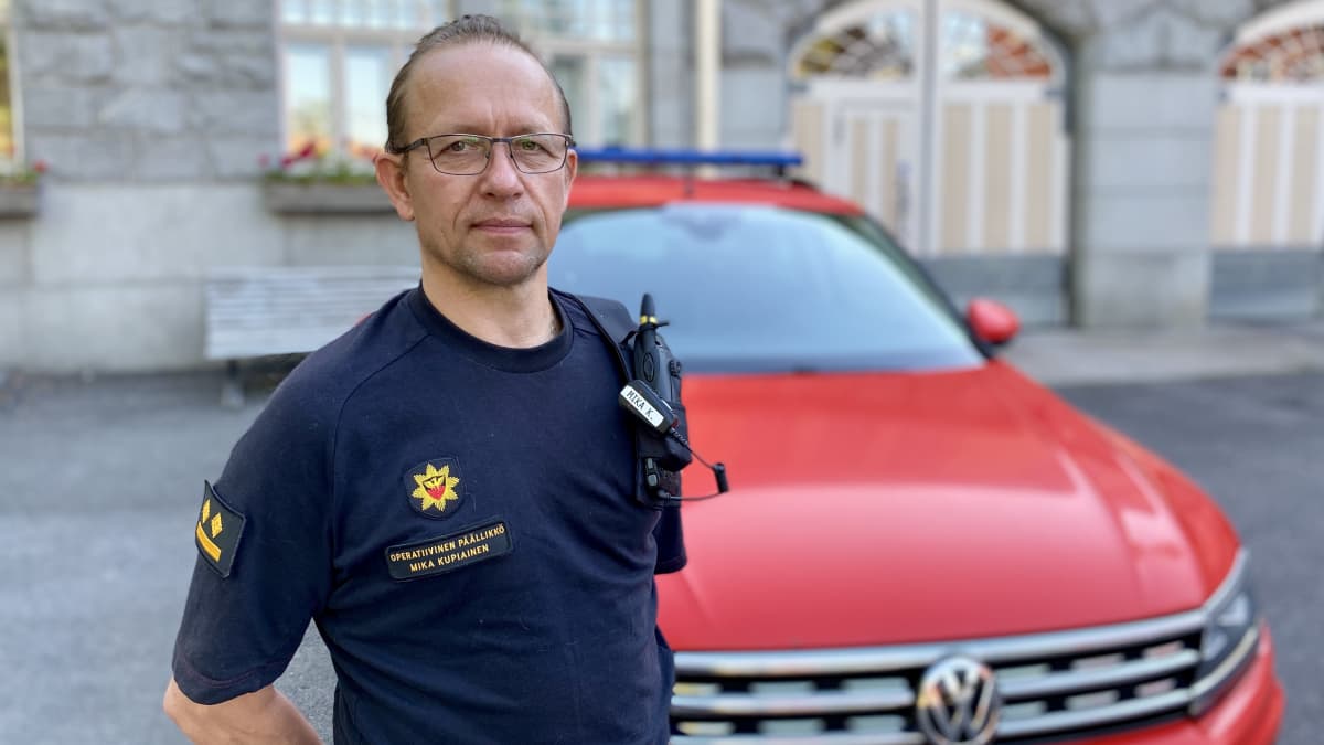Päivystävä palopäällikkö Mika Kupiainen seisoo paloaseman pihalla. Takana punainen auto. 
