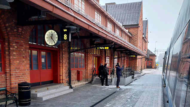 Three people on a platform with a red-brick station to the left and a silver train to the right.
