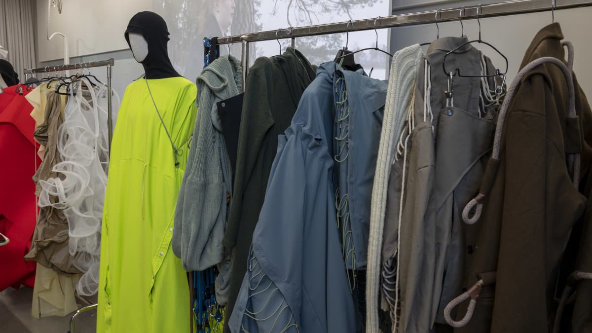 Mallinuken päällä neonkeltainen koko vartalo mekko ja vieressä vaatetelineissä roikuu murrettua värisarjaa olevia takkeja, joissa on paljon naruja ja köysiä kiinnitettyinä.