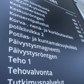 Musta sairaalan poliklinikkojen ja palvelupisteiden opastuskyltti, jossa valkoisella tekstillä eri osastojen nimiä ja lyhenteitä.