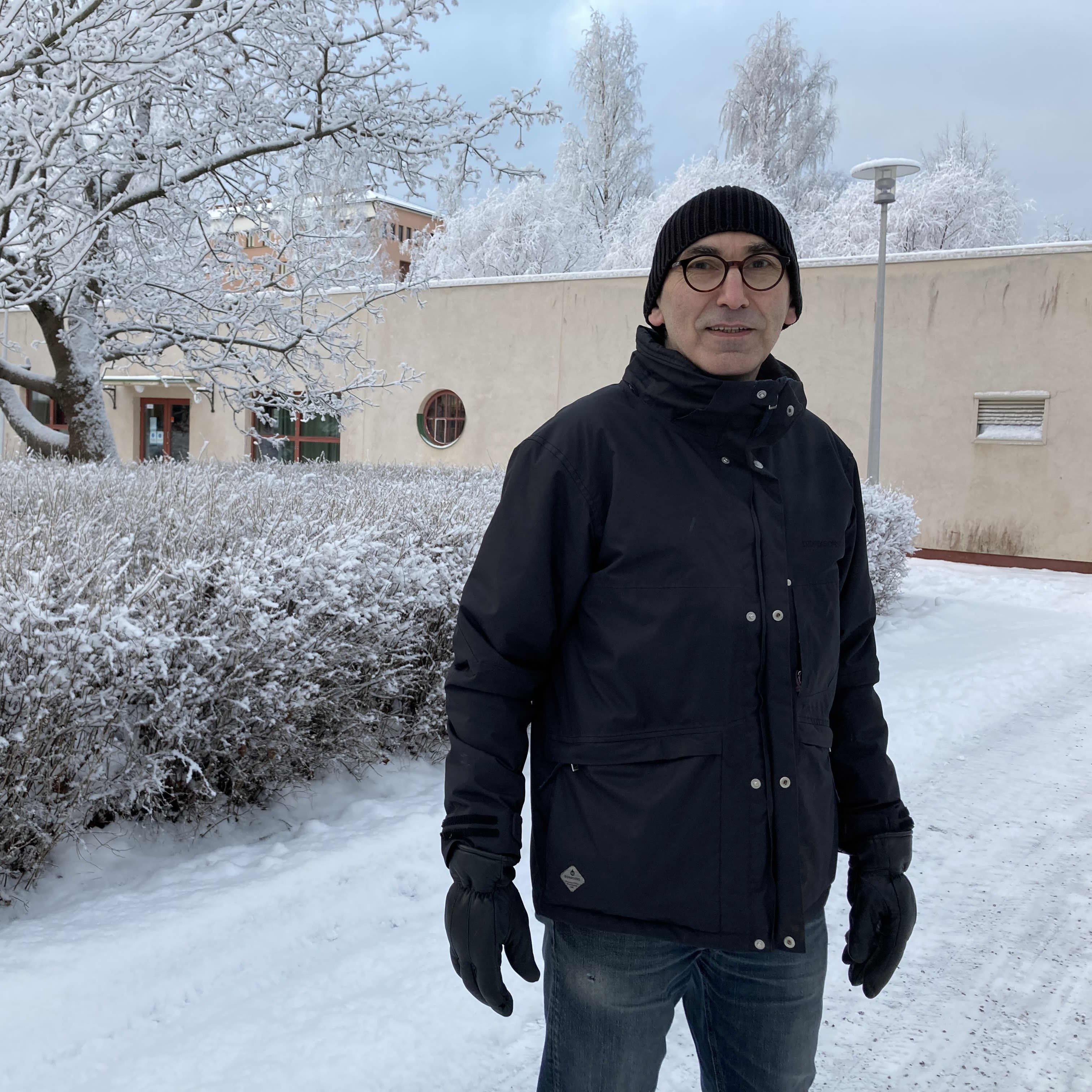 Professori Juha T. Hakala seisoo lumisessa maisemassa pensasaidan edessä.