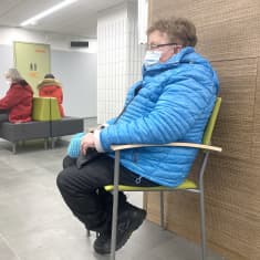 Kolme asiakasta istuu Harjun terveydn käytävällä odottamassa vuoroaan. Etualan naiselle sininen untuvatakki, kauempana kaaksi naista istuu sohvalla punaisissa takeissaan.