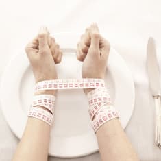 Naisen kädet tyhjällä lautasella ja kädet sidottu yhteen mittanauhalla.