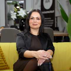 Tummatukkainen nainen katsoo kameraan. Hän istuu keltaisella sohvalla.