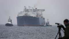 Valtava LNG-alus (283 metriä pitkä) lipuu satamaan, kaksi pienempää alusta saattelee sitä. Etualalla pipopäinen mies kuvaa aluksen saapumista sumuisella säällä.