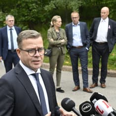Petteri Orpo speaks in front of microphones outdoors while three men in dark suits and a woman in a light-brown suit stand in the background, with grass and bushes behind them.