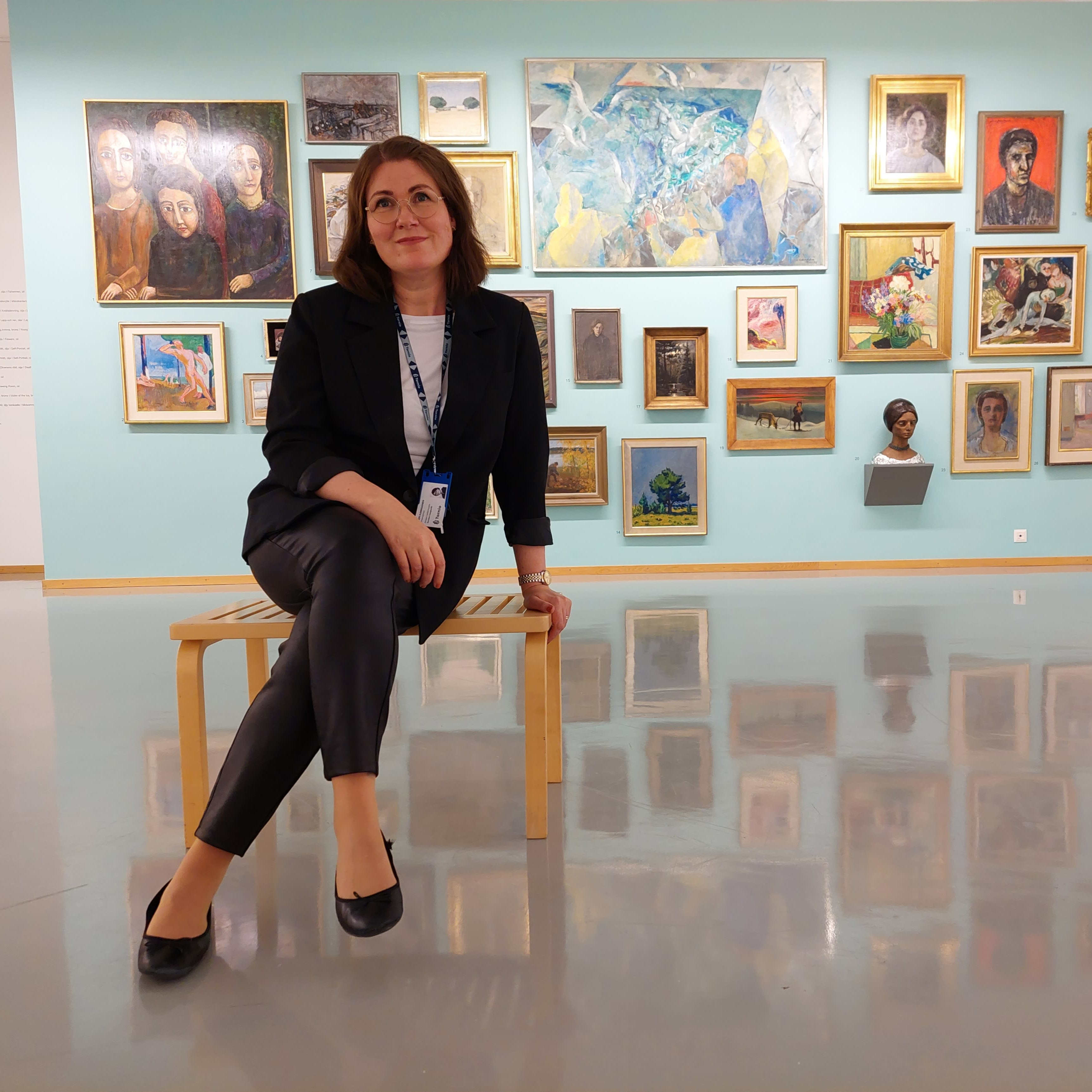 Aineen taidemuseon uusi johtaja Virpi Kanniainen istuu näyttelysalissa penkillä. Taustaseinällä on värikkäitä maalauksia ja veistoksia.