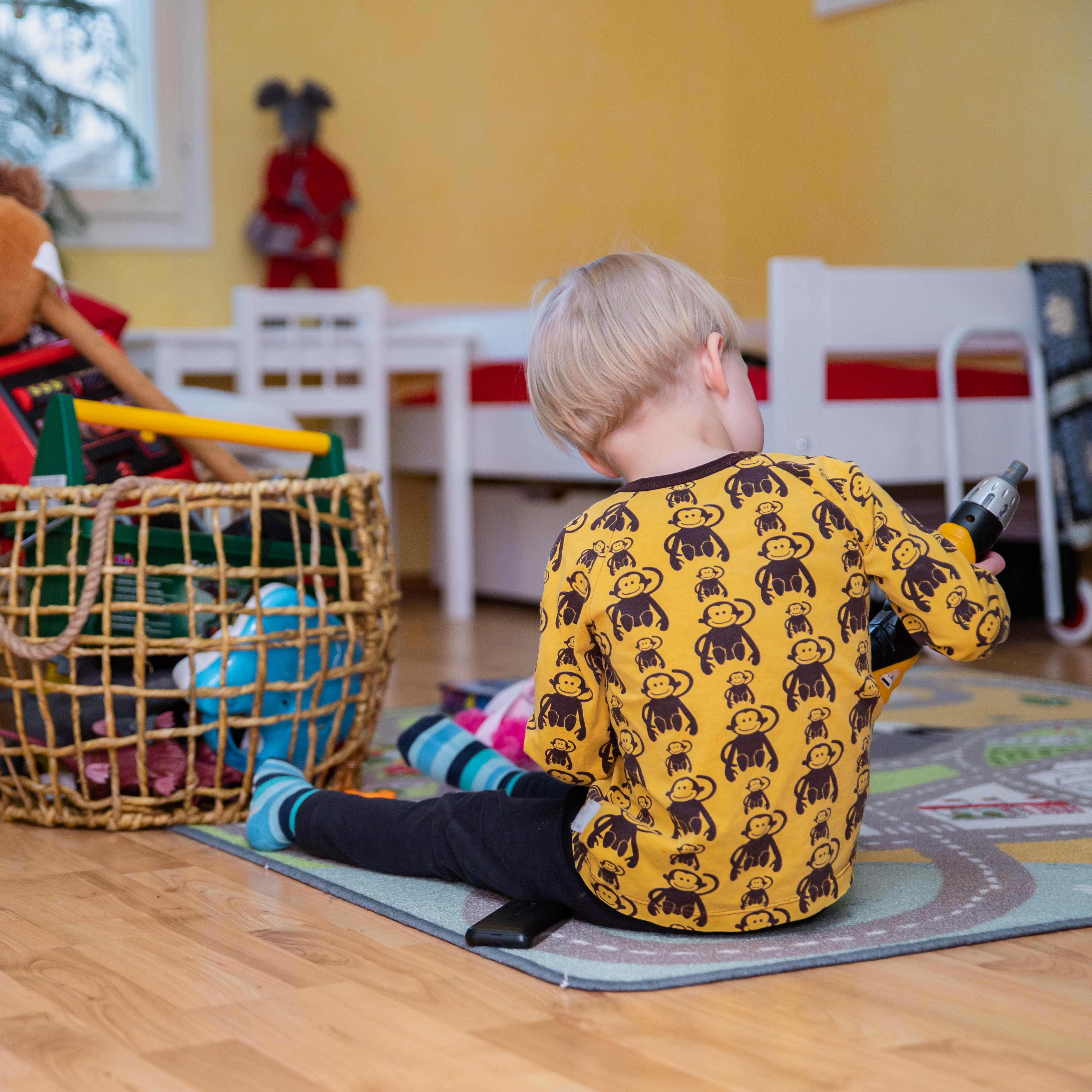 Lapsi leikkii leluilla lattialla istuen. Kädessä on lelupora. Lapsen vieressä lelukori.