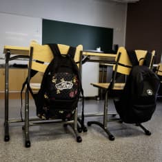 Kaksi reppua roikkuu tuolien selkänojissa tyhjässä luokkahuoneessa. 